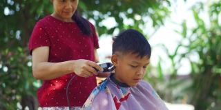 亚洲母亲正在修剪她的儿子的头发在早上在花园里。在COVID-19或冠状病毒大流行期间的国内