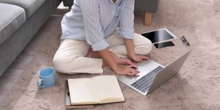 一位年轻的亚洲女商人正在笔记本电脑上寻找信息，她在笔记本上做笔记，她坐在家里客厅的地毯上。商业女性和工作理念。