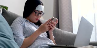 年轻的亚洲女商人感到头痛和发烧。她在家里客厅的沙发上用智能手机和笔记本电脑工作。在家工作，商业女性和工作理念。