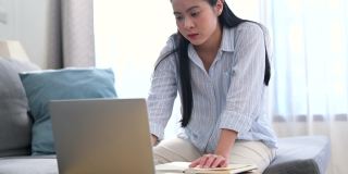年轻的亚洲女商人正在笔记本电脑上寻找信息，她在笔记本上做笔记，她坐在家里客厅的沙发上。商业女性和工作理念。