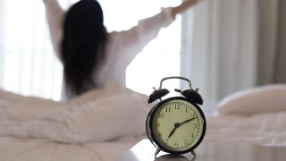 特写闹钟有一个美好的一天与背景快乐的女人伸展在床上醒来后，阳光在早晨。视频素材模板下载