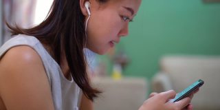 年轻的亚洲女性戴着入耳式耳机，使用智能手机