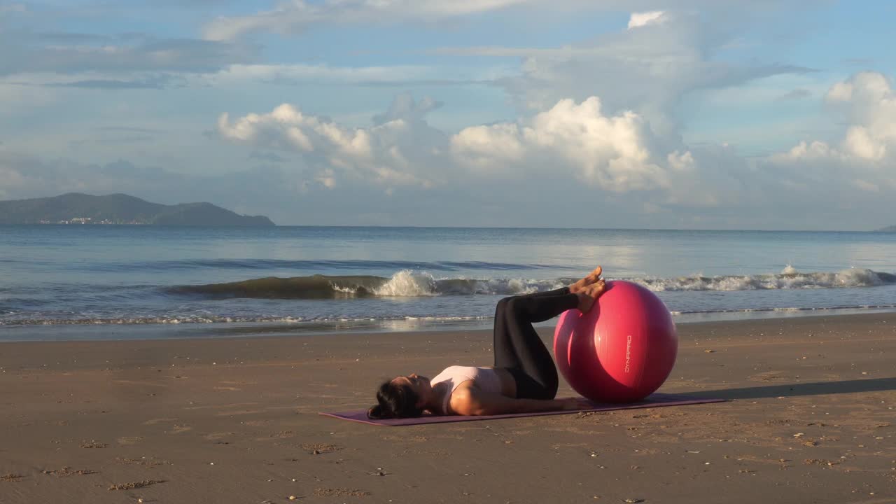 健康的亚洲美女早上在海滩上用球做瑜伽练习。