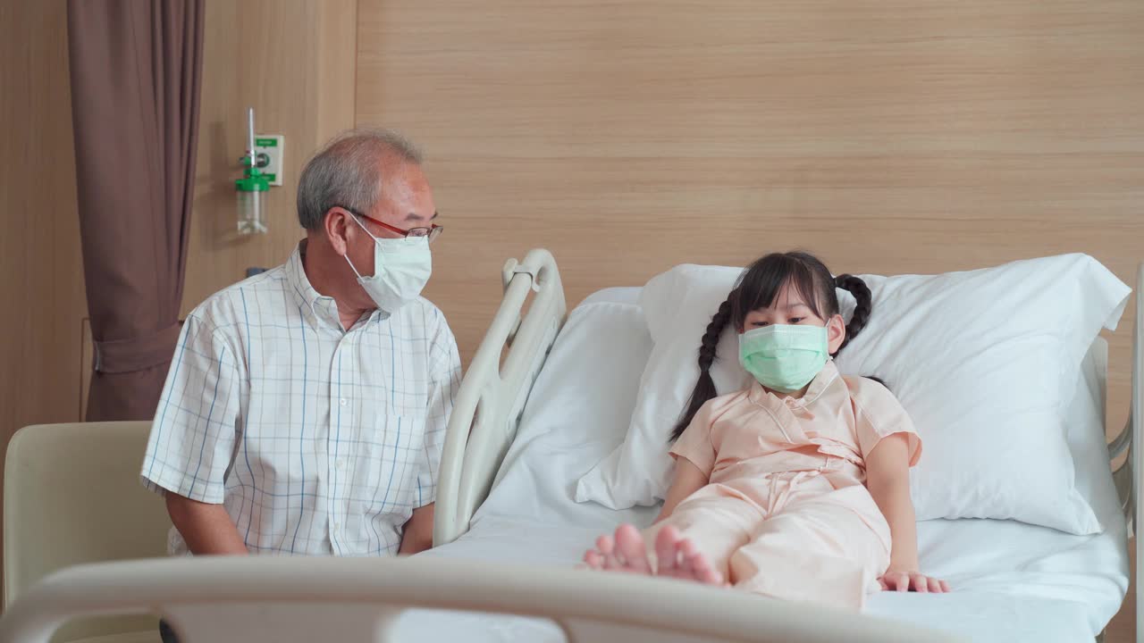 亚洲男医生带着口罩来到康复室，向躺在病床上的儿童病人询问健康问题。男医生检查心率、脉搏和提供健康保健咨询。