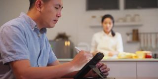 亚洲父亲写便条和工作平板电脑等待他的妻子和女儿准备晚餐的食物。他很忙，在新冠肺炎大流行期间不得不在家工作。新标准的概念。