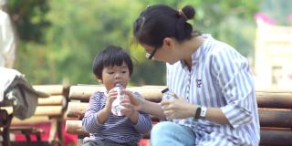 亚洲母亲和她的儿子坐在公园的长凳上喝水