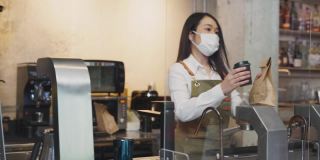 在餐厅柜台，穿着白色制服的亚洲女服务员将棕色的空白纸袋放在压克力隔板之间递送给顾客，两人都戴着口罩。2019年冠状病毒大流行。