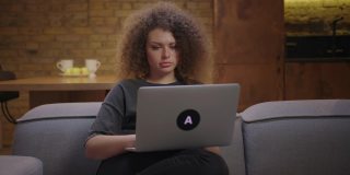 20多岁的卷发女人坐在家里的沙发上用笔记本电脑工作。年轻专注的女性在膝上的笔记本电脑上打字。