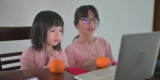 亚洲的中国小女孩和她的妹妹看着笔记本电脑的摄像头，用红包和橘子向她的祖父母祝贺春节