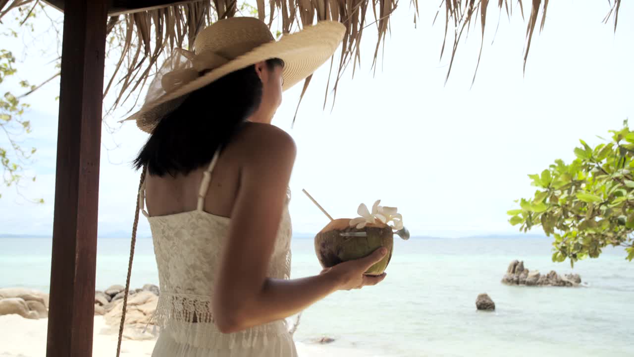美丽年轻的亚洲女人喝椰子汁沿着海滩。