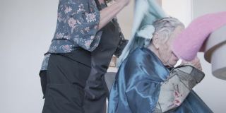 手持拍摄的专业护理发型师干燥一个年长的白人妇女的白色和灰色的头发在厨房里在一个洗发集在家