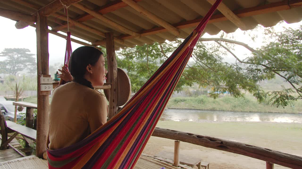 亚洲美丽的女人坐在阳台上的度假酒店在热带森林背景周末度假。快乐独自旅行的概念。4 k慢动作。