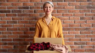 一位面带微笑的资深亚洲女志愿者拿着一个纸板箱，里面装着捐赠的衣服，用于旧衣服的再利用。一个快乐的女人的肖像靠着捐款箱站在砖墙上视频素材模板下载