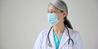 特写视频肖像的亚洲灰头发女医生在医疗制服，防护口罩和听诊器站在白色的背景下，看着镜头。医疗保健的概念