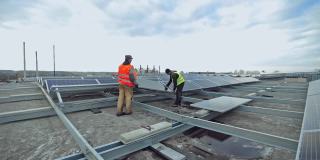 技术人员在金属底座上安装太阳能电池板。两名工人正在平屋顶上安装太阳能电池板。光伏板将太阳能转化为电能。