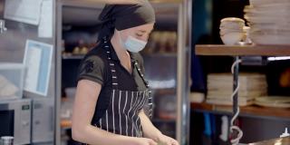 穿着围裙和Covid-19口罩的年轻女厨师正在为餐厅的冰箱准备材料。一名在厨房工作的严肃白人女性正在应对冠状病毒大流行。