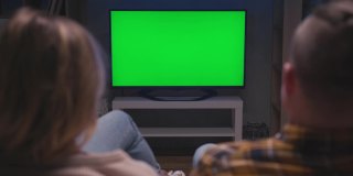 一对家庭夫妇一起坐在客厅的沙发上看绿屏电视模型。国内电影中看绿屏电视的休闲人士的后视观。在家休息看电视节目或新闻