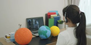 一名亚洲少女通过客厅的笔记本电脑屏幕向老师在线展示她的科学作品，当时学校因COVID-19疫情而停课。