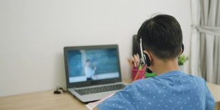 一名亚洲男孩在学校因新冠肺炎疫情关闭期间，通过笔记本电脑与老师在线学习数学，并在家里的客厅里看摄像机。