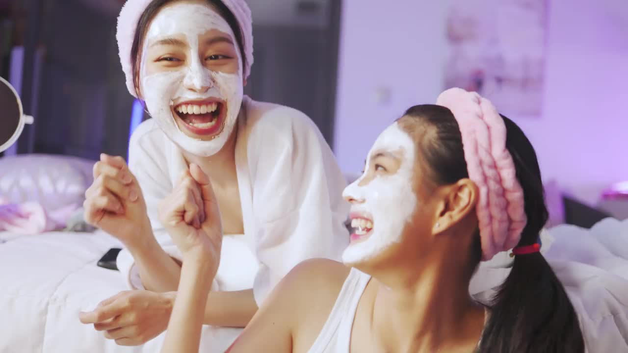 亚洲两个年轻的女人正在放松水疗中心在家里一起笑放松，微笑的女孩朋友有乐趣。虚拟活动，身体护理在家。年轻女子敷面膜。水疗中心提供各种保健服务。