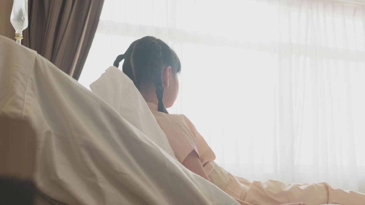 亚洲小女孩戴着口罩躺在康复室的床上，等待家人的探望。父亲微笑着，幸福地拥抱着女儿。