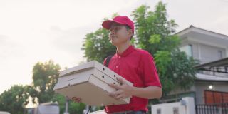 亚洲送货男工人穿着红色制服搬运食物，披萨给年轻漂亮的女顾客在房子前面。邮差和快递杂货配送服务理念