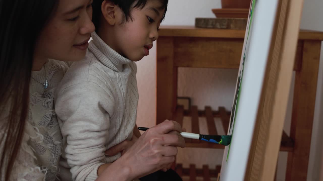 亚洲母亲有乐趣做颜色油漆与小儿子在画布上在家-母亲和孩子的爱