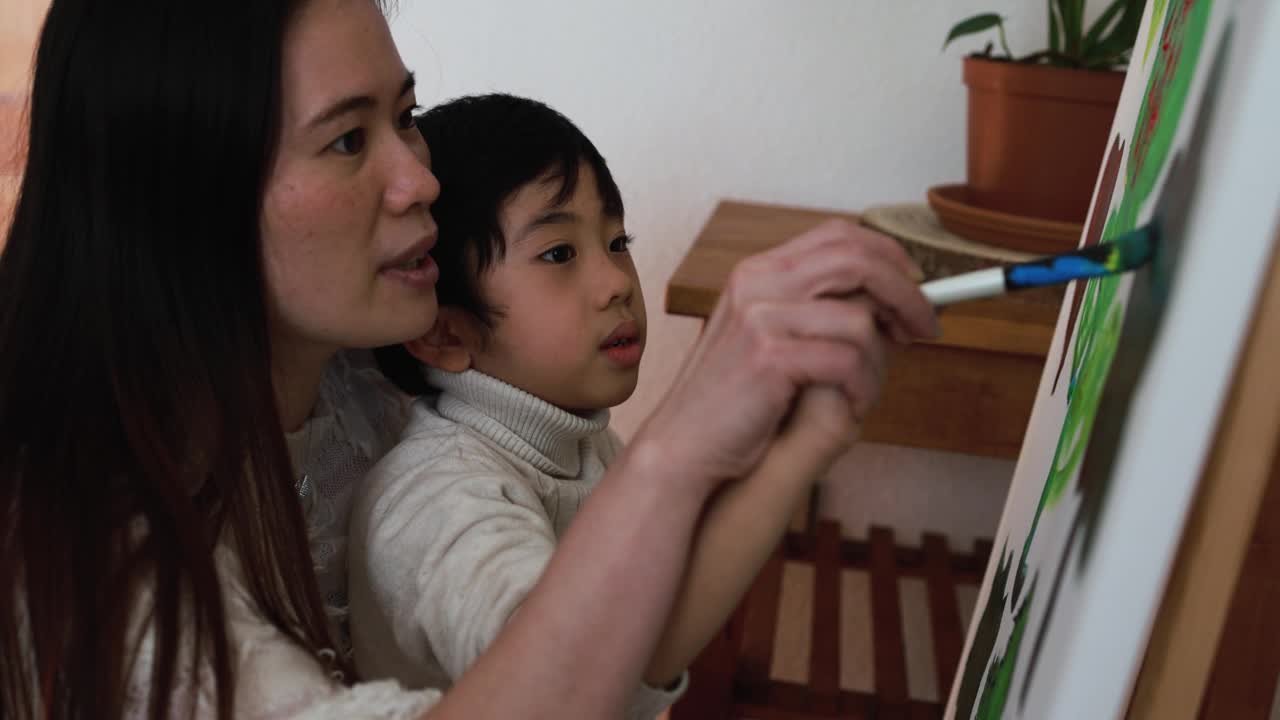 亚洲母亲有乐趣做颜色油漆与小儿子在画布上在家-母亲和孩子的爱