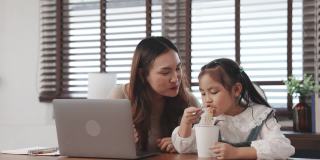 自由职业者亚洲母亲工作在桌子上与她的笔记本电脑，她的女儿吃面条在客厅在家里，幸福的家庭人的生活方式休闲活动概念。