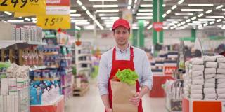 穿着红色制服的售货员拿着装着新鲜蔬菜的牛皮纸包站在超市里。