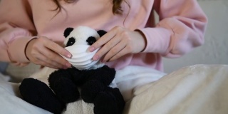 一个女孩正在给一个熊猫玩具戴上口罩，以防止冠状病毒感染