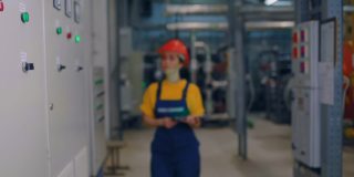 一位年轻的女工程师戴着防护头盔，手里拿着平板电脑，微笑着走向相机。现代工业和生产