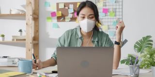 亚洲女商人戴着医用口罩，使用笔记本电脑与同事在视频通话中讨论计划，同时在家起居室工作。