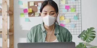 亚洲女商人戴着医用口罩，使用笔记本电脑与同事在视频通话中讨论计划，同时在家起居室工作。