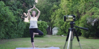 亚洲女性在线教练瑜伽示范通过移动智能手机三脚架直播观众的姿势。女性健身辅导。教练与在草地上在线教授瑜伽的学生交谈。