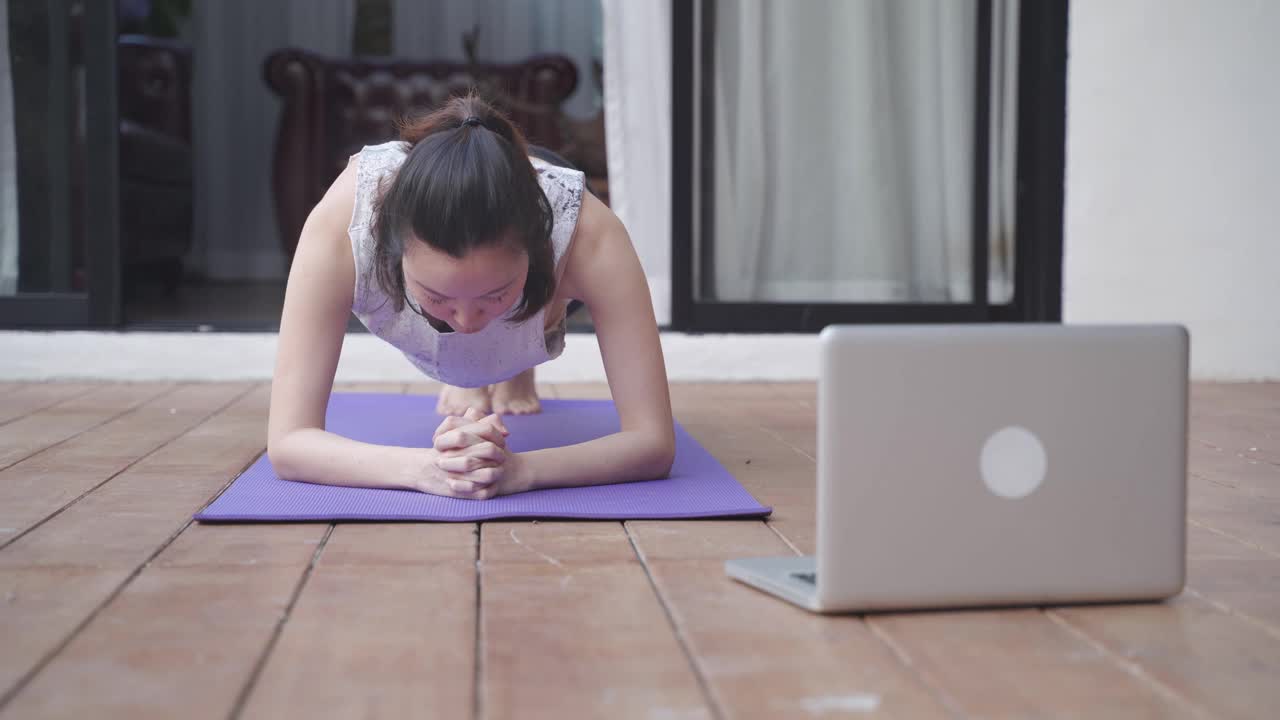 亚洲女人做瑜伽姿势锻炼在阳台上在家。女性在电脑上看在线教程。锻炼伸展莲花和平板支撑姿势。