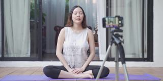 亚洲女性在线教练瑜伽示范通过移动智能手机三脚架直播观众的姿势。女性健身辅导。教练与在家里在线教授瑜伽的学生交谈。