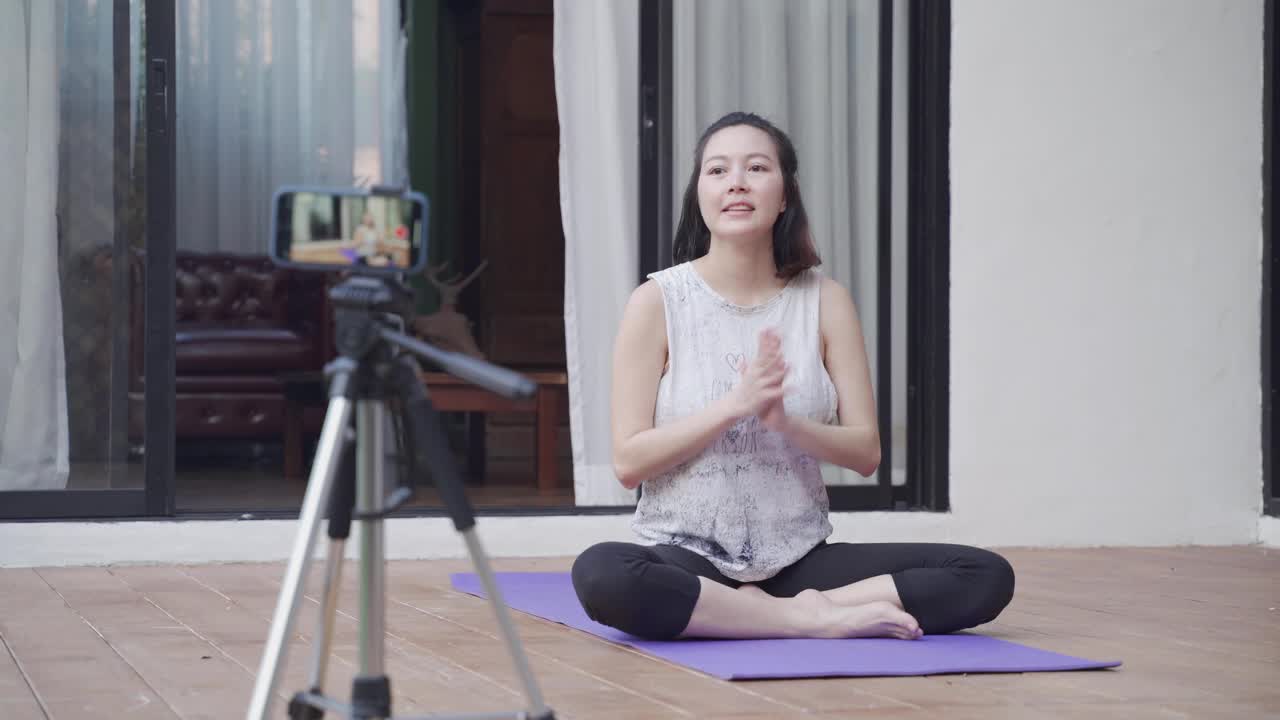 亚洲女性在线教练瑜伽示范通过移动智能手机三脚架直播观众的姿势。女性健身辅导。教练与在家里在线教授瑜伽的学生交谈。