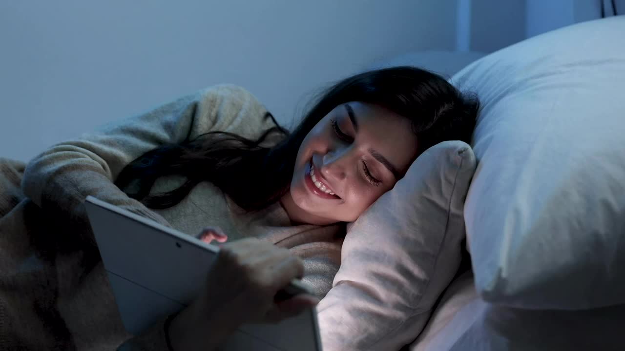 亚洲女性在晚上睡觉前在床上使用平板电脑，手机成瘾概念，蓝光对眼睛有害。