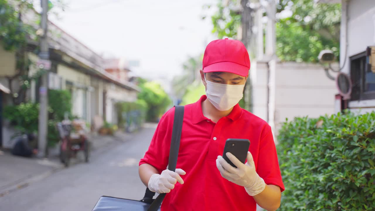 在冠状病毒大流行期间，身穿红色制服、戴面罩的亚洲快递员携带食品袋，将食品送到顾客家中。他走在村外寻找客户的家庭地址。