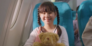亚洲小女孩拥抱熊娃娃的肖像，坐在飞机上。孩子感到兴奋和快乐的旅行与她的家人。乘客在飞机上的运输概念。