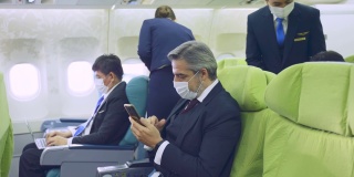 在新冠肺炎大流行期间，白人商人在飞机上戴口罩使用手机，以防止感染新冠肺炎。该男子正在等待机上乘务员提供的机上服务。