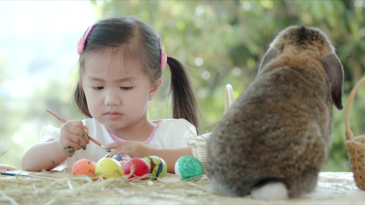 亚洲小女孩在复活节书桌上的兔子旁边给复活节蛋涂色。