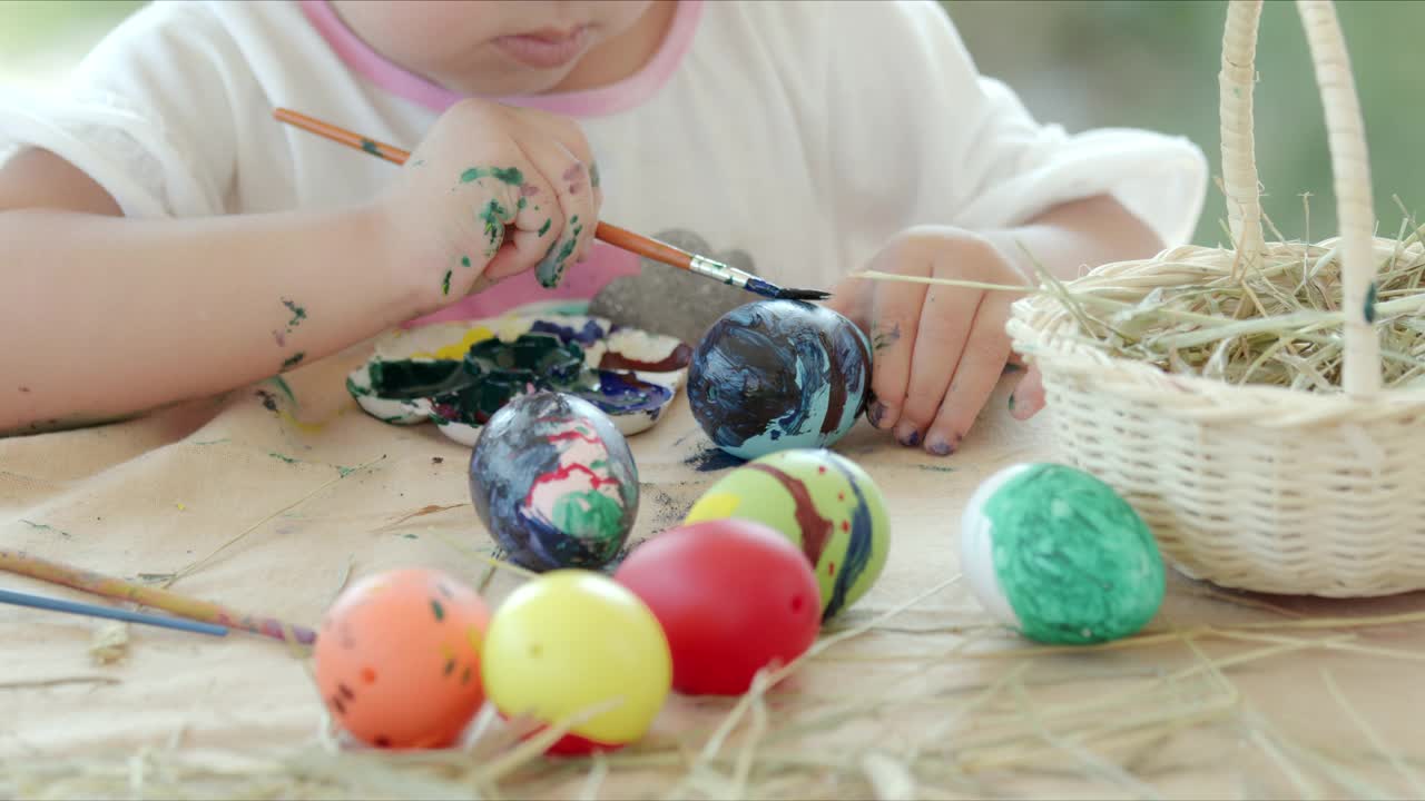 小女孩在书桌上用画笔和颜料给复活节彩蛋涂色。