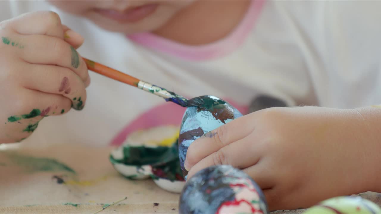 小女孩在书桌上用画笔和颜料给复活节彩蛋涂色。