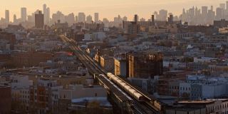 从住宅区远眺布鲁克林市中心和曼哈顿。傍晚，一列火车沿着默特尔大道沿着高架地铁开往布鲁克林市中心。无人机视频与前摇摄像机运动。