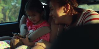 可爱的亚洲小女孩坐在汽车座椅上，和她年轻的妈妈一起在车里看智能手机里的卡通。