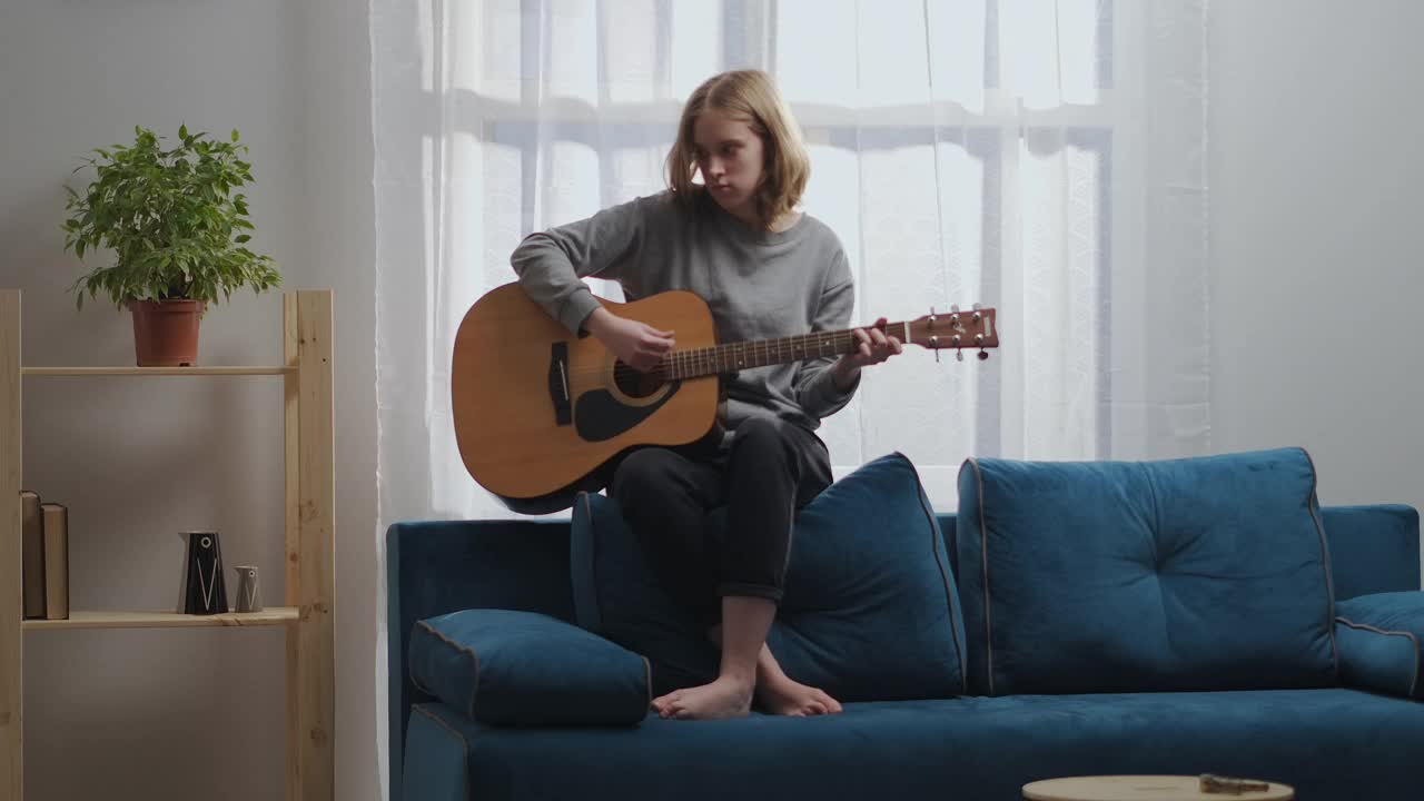 一个穿着灰色运动衫和黑色裤子的少女在吉他上弹奏着抒情的旋律。坐在蓝色沙发的背面。太阳从后面的窗户射进来。