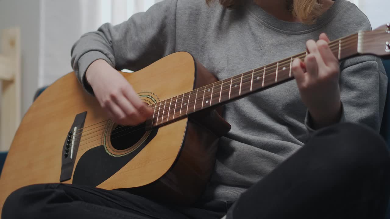 一个穿着灰色运动衫的少年在吉他上弹奏着一首抒情的旋律。以舒适的莲花姿势坐在一个蓝色的沙发上。阳光从窗户射进来。近距离拍摄