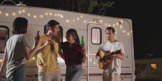 一群亚洲男女朋友在晚上的户外聚会上一起弹吉他。朋友们带着幸福和微笑碰杯喝啤酒。乘露营车旅行大自然。
